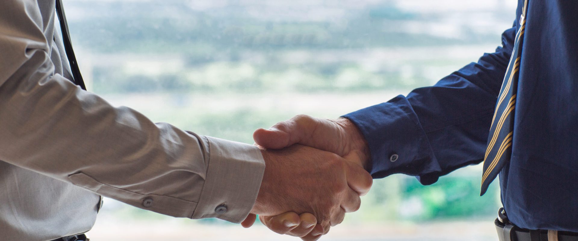 Closeup of two businessmen shaking hands with blurry city view in background. People are seen partly.